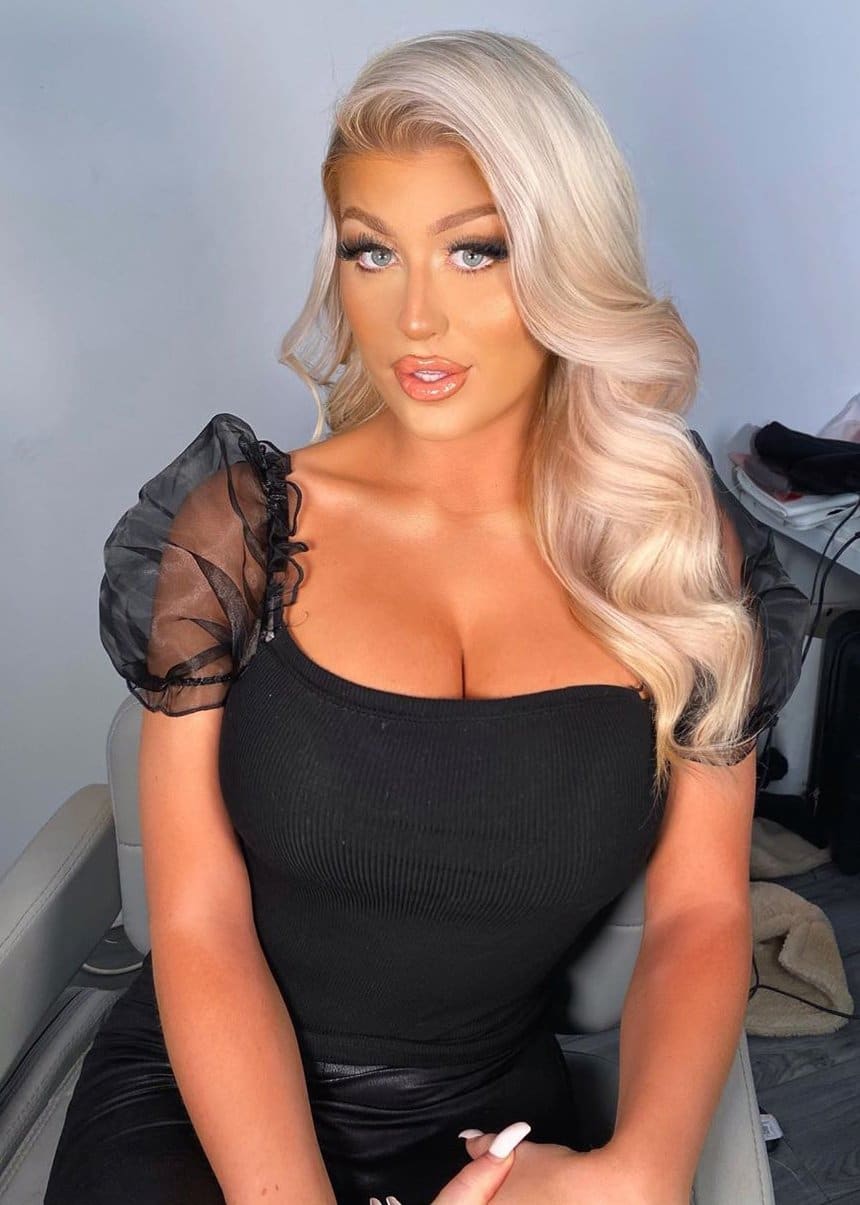 Eva sits facing forward in a light chair, wearing a black ribbed top with sheer puffed sleeves and a low square neckline against a plain wall.