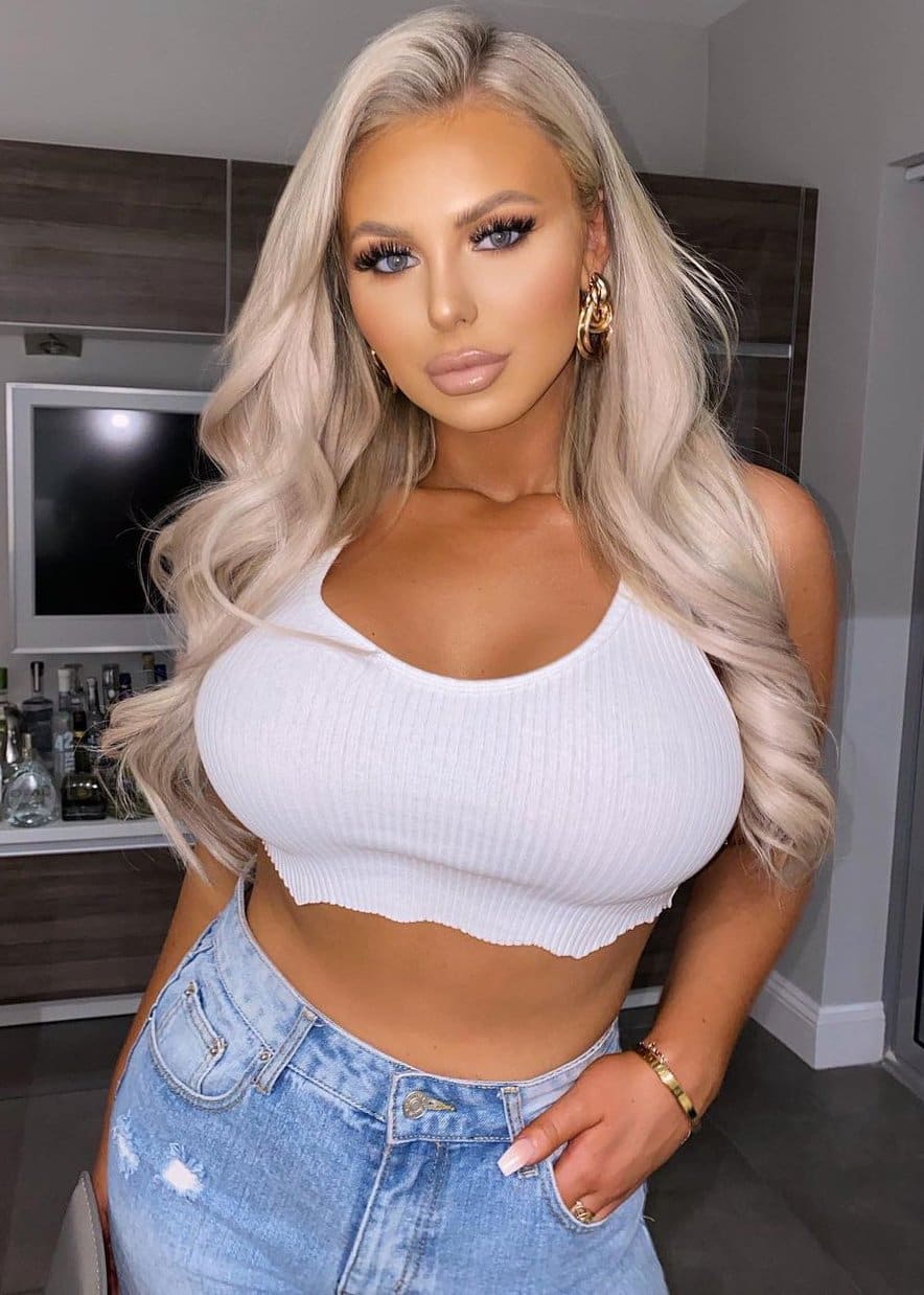 A woman in a white ribbed crop top and distressed jeans stands in a room with a TV and shelf of bottles behind her.