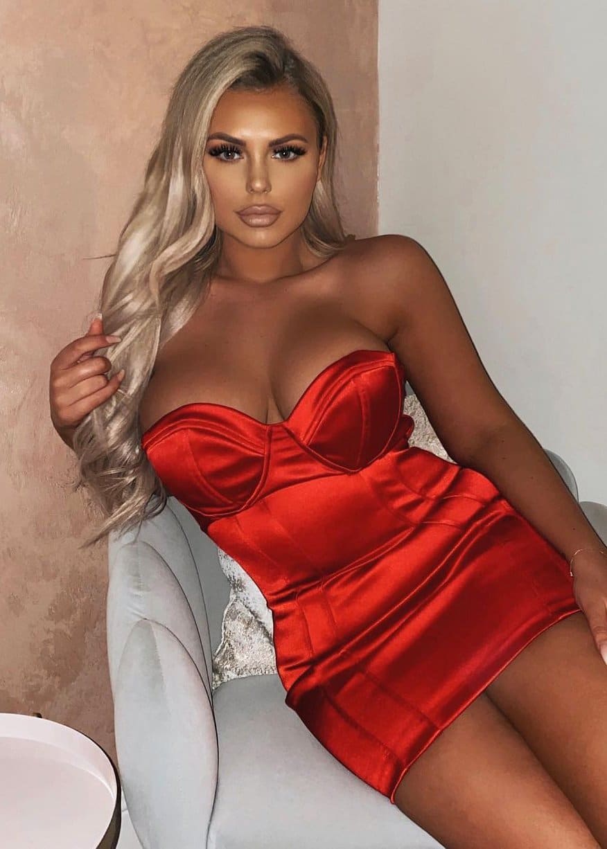 A woman sits on a light chair in a strapless red satin mini dress, touching her hair against a textured beige wall.