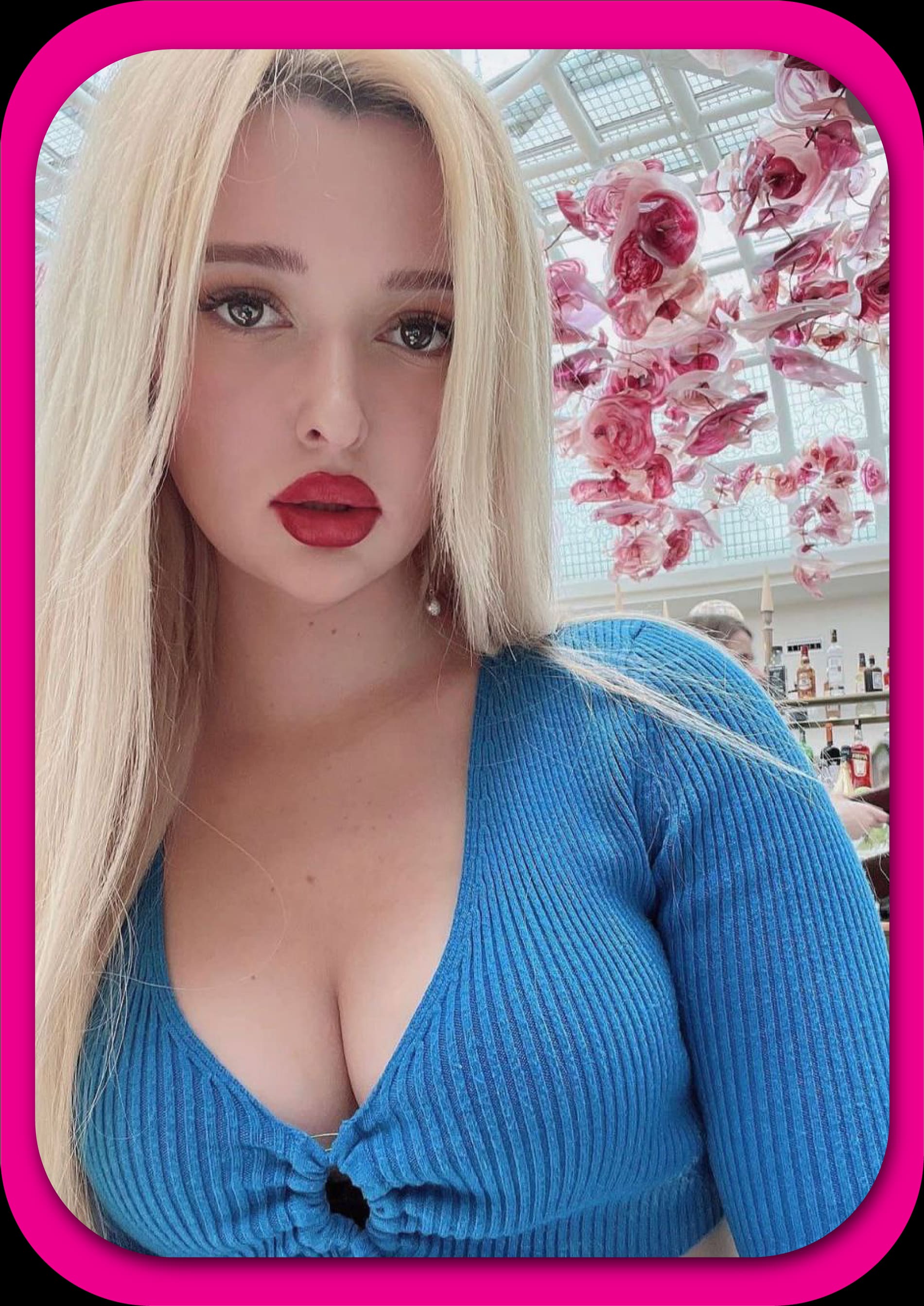 A blonde woman in a blue ribbed V-neck top poses in a close-up selfie near pink floral decorations and a bar area.