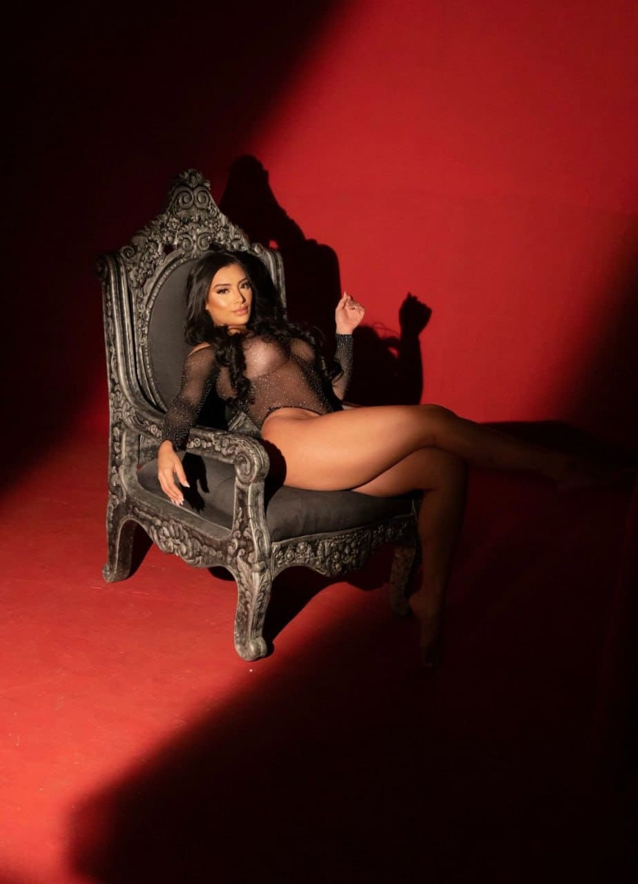 A woman in a sheer black bodysuit sits on an ornate chair against a red wall, her bare legs crossed in the frame.
