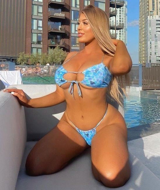A blonde woman kneels on a ledge beside a pool in a blue patterned bikini, touching her hair, with high-rise apartment buildings in the background.