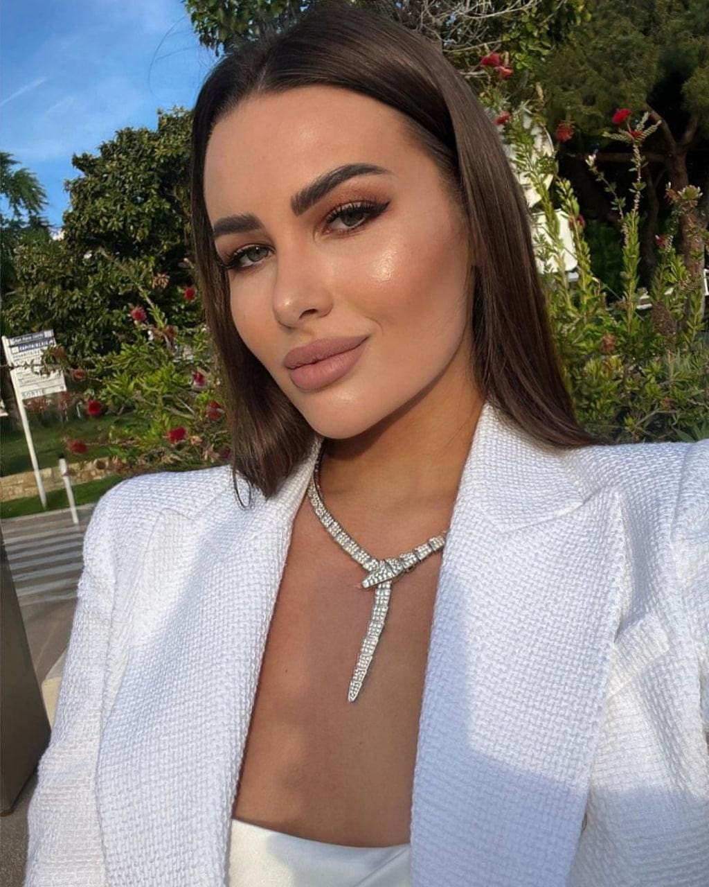 Kimberly poses outdoors in a white textured blazer over a white top, wearing a large diamond necklace against a backdrop of greenery and blue sky.