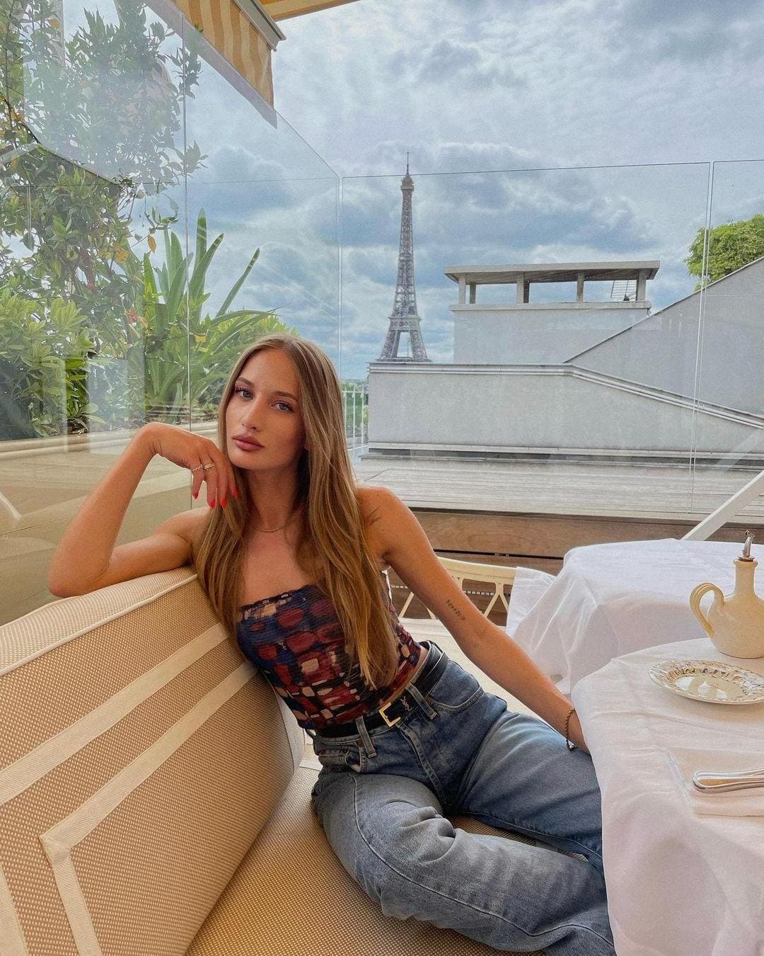 April sits on a woven terrace bench in a strapless patterned top and blue jeans, with the Eiffel Tower visible behind her under a cloudy sky.