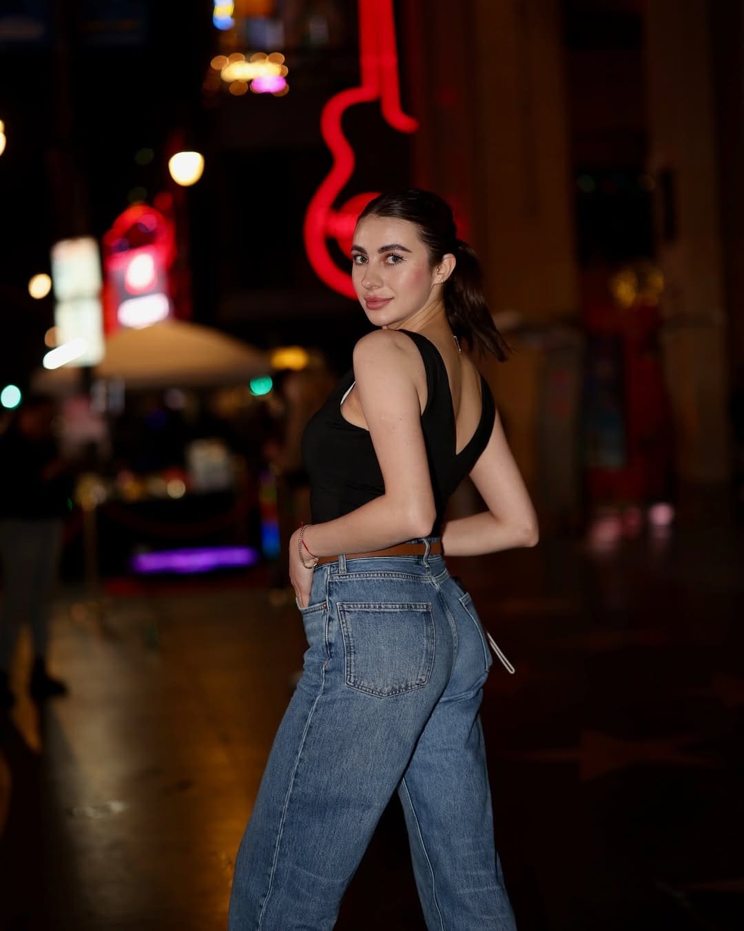 A woman with a dark ponytail stands in profile on a city street at night, wearing a black sleeveless top and light blue jeans.