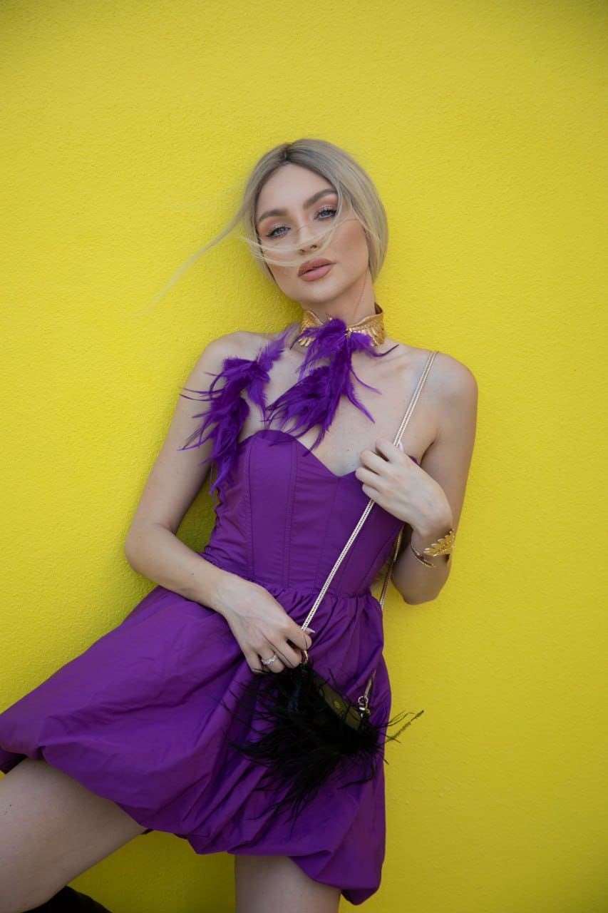 A blonde woman with wind-blown hair wears a purple feathered mini dress and holds a black handbag against a yellow wall.