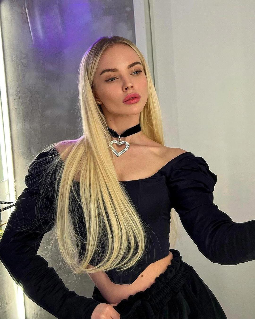 Standing indoors, she wears a black square-neck top and heart pendant choker, hand on hip, against a wall with purple glow.
