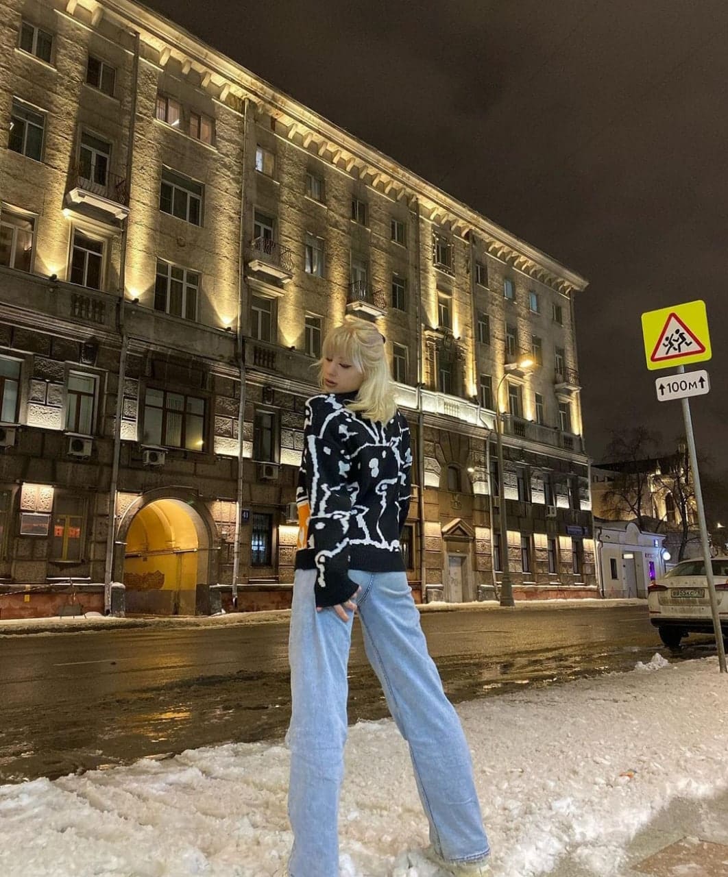 Woman in patterned turtleneck and wide-leg jeans stands on snowy street at night near lit building.