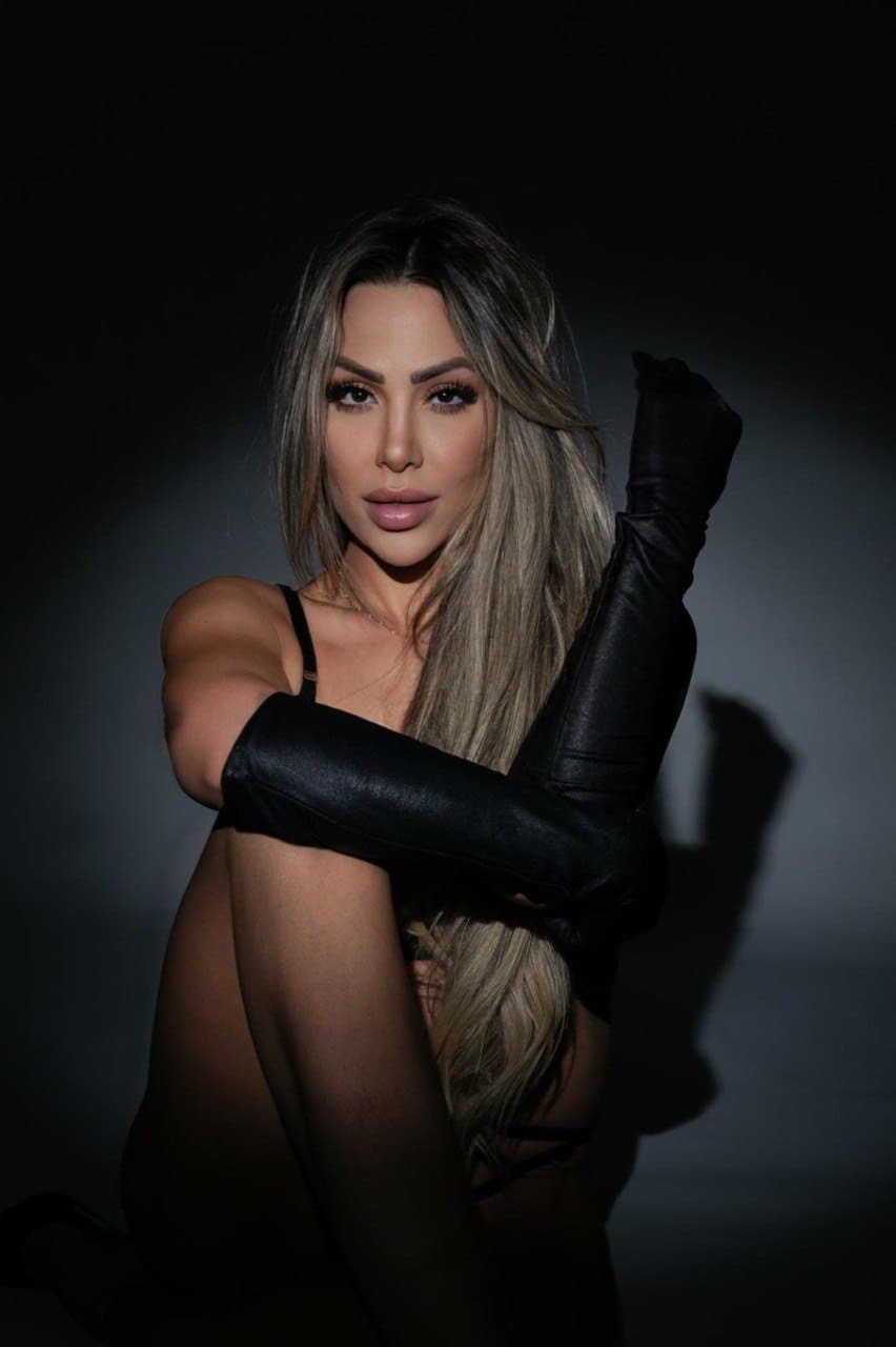 Seated against a dark backdrop, a woman in leather thigh-high boots and a thin-strapped top crosses her legs.