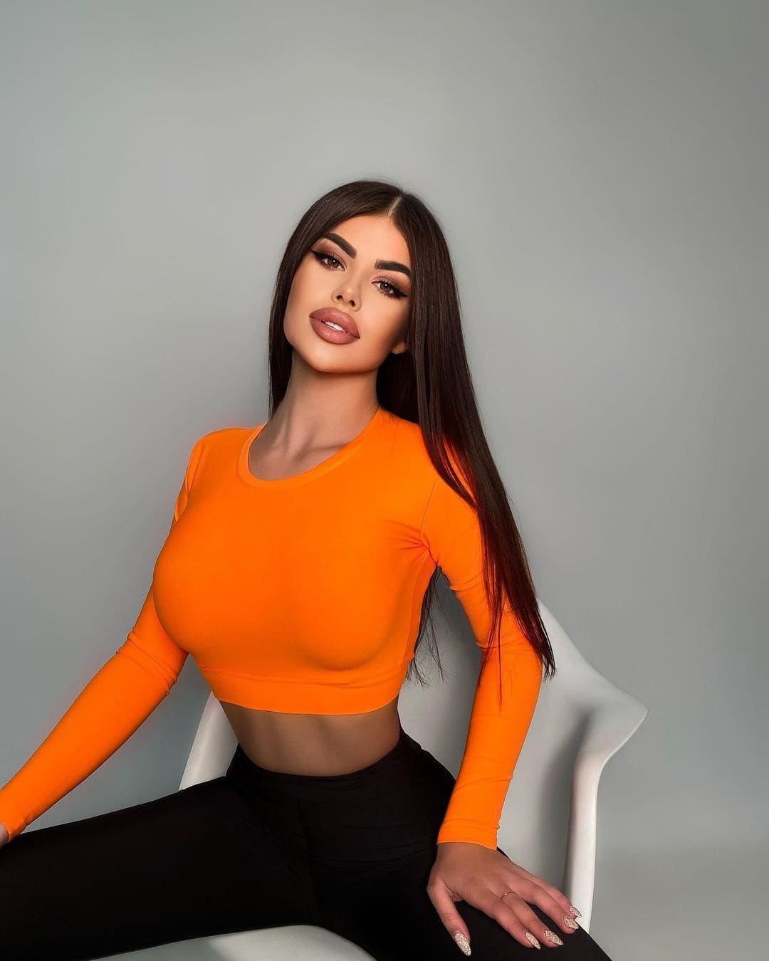 Lyra sits on a white chair against a gray backdrop, wearing an orange crop top and black pants with her midriff exposed.