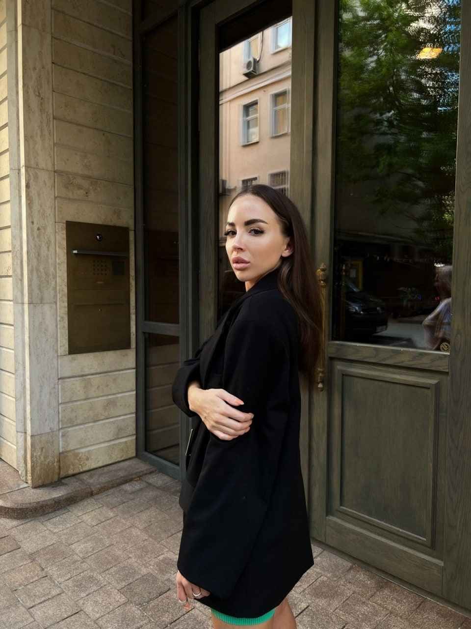 A woman stands before a glass door with arms crossed, wearing a black blazer over a green top, looking toward the camera.