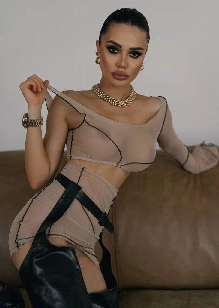 She sits on a brown couch wearing a sheer beige two-piece outfit with black trim, a gold chain necklace, and black leather thigh-high boots, holding a top strap.