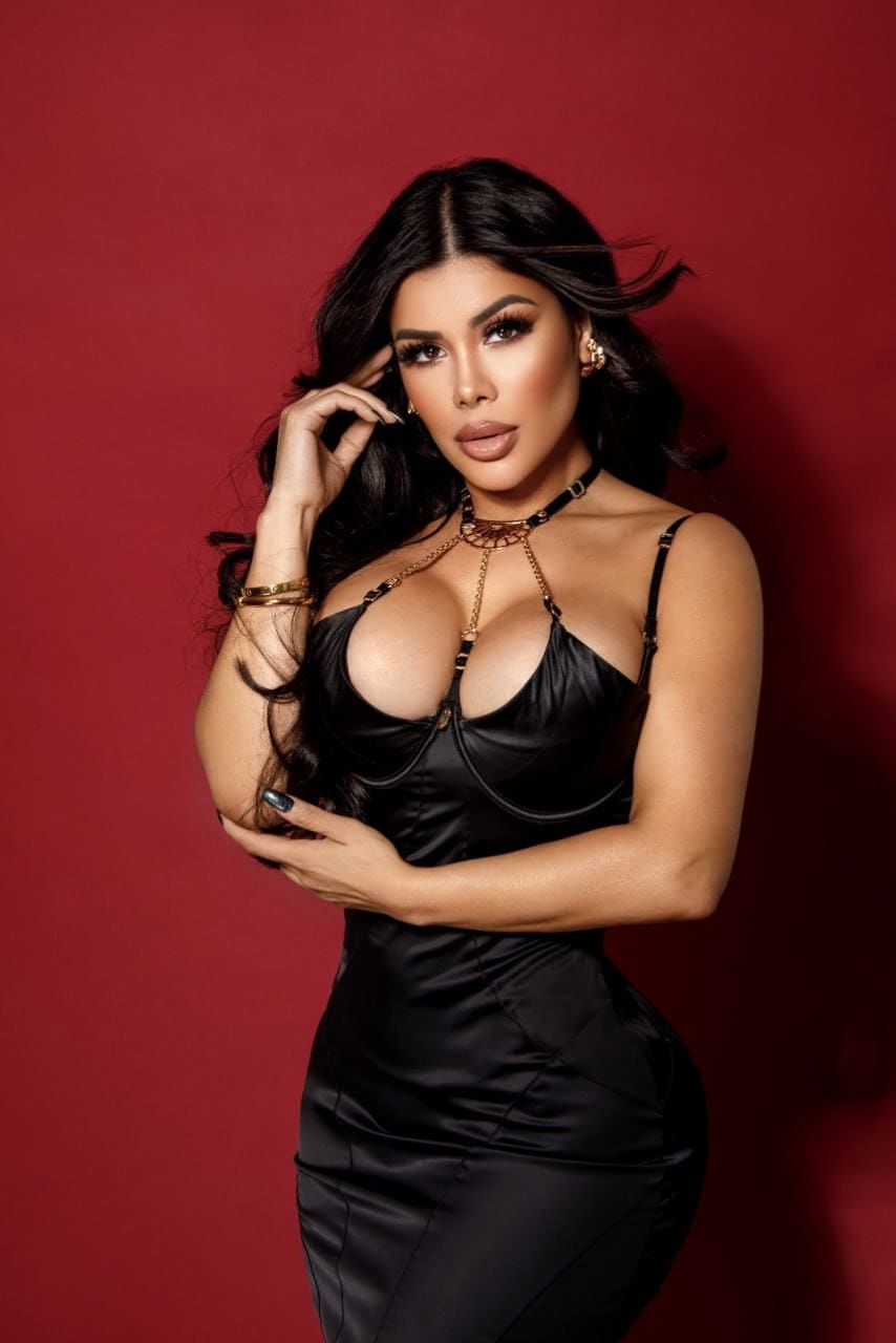 Camila stands against a red backdrop in a black satin slip dress, one hand on her chest and the other touching her dark wavy hair.