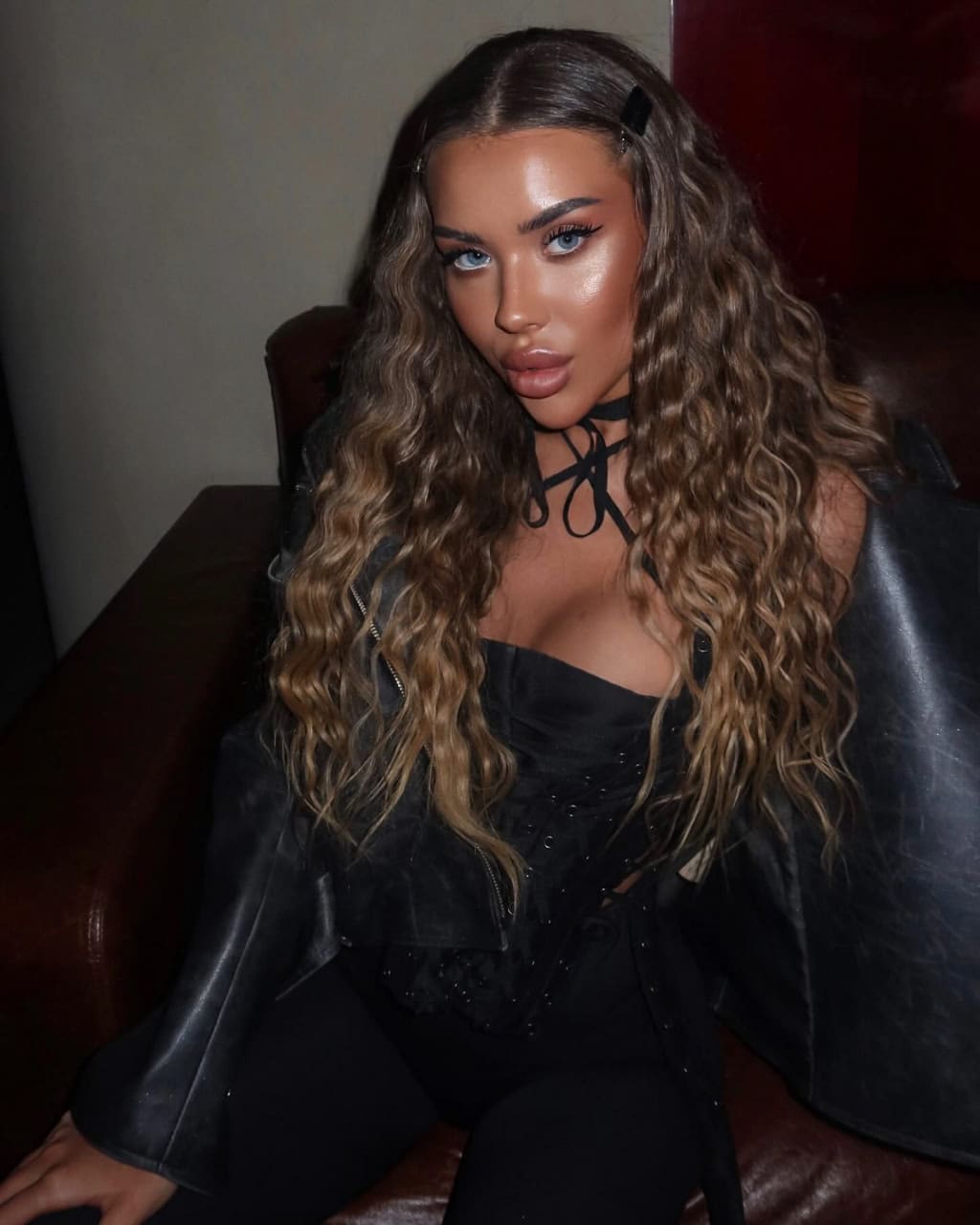 A woman sits on a dark leather couch in a dimly lit room, wearing a black leather jacket over a low-cut top and a choker.